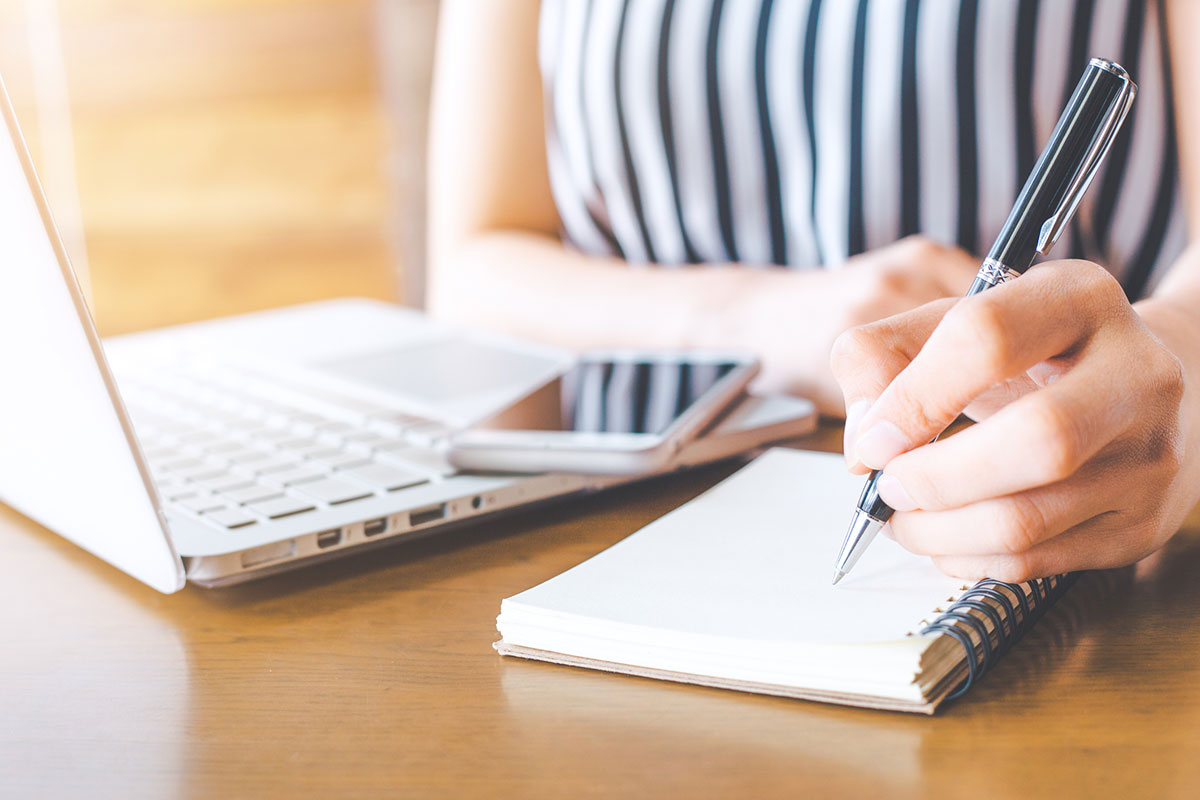 hands of person at work with a laptop open and a paper notepad, taking notes with a pen