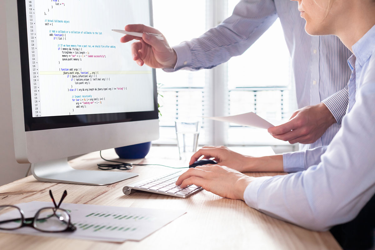 two web developers talking while one is coding via a monitor and keyboard