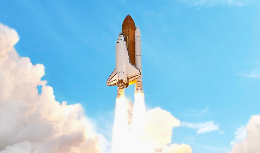 space shuttle rockets firing, launching into a clear, blue sky with billows of smoke and clouds below 