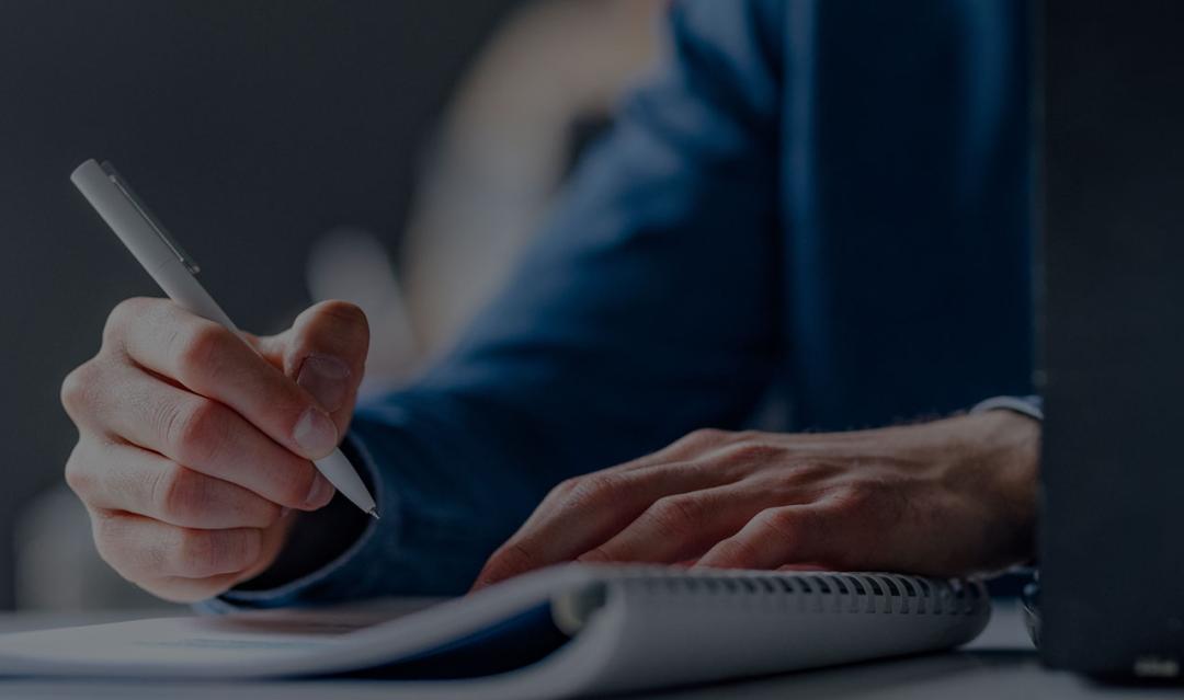 man taking notes with pen and paper, laptop is open 