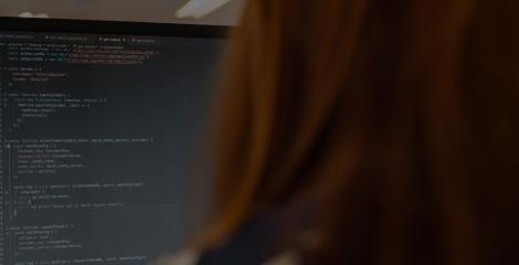 backside view of a person with long red hair looking at a computer screen and code