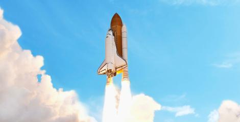 space shuttle rockets firing, launching into a clear, blue sky with billows of smoke and clouds below 