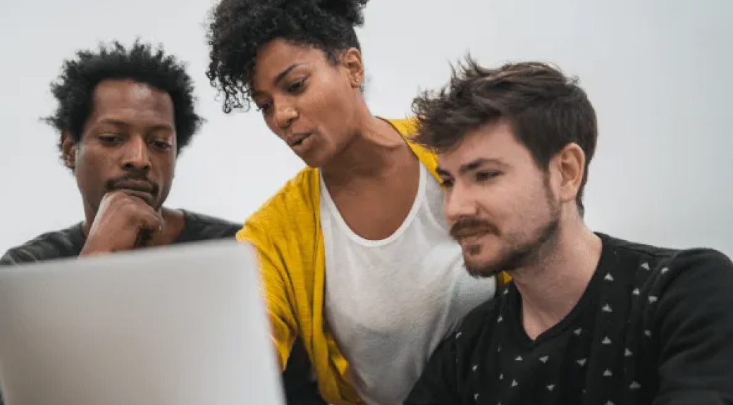 woman in yellow cardigan giving help to two men seated around a computer