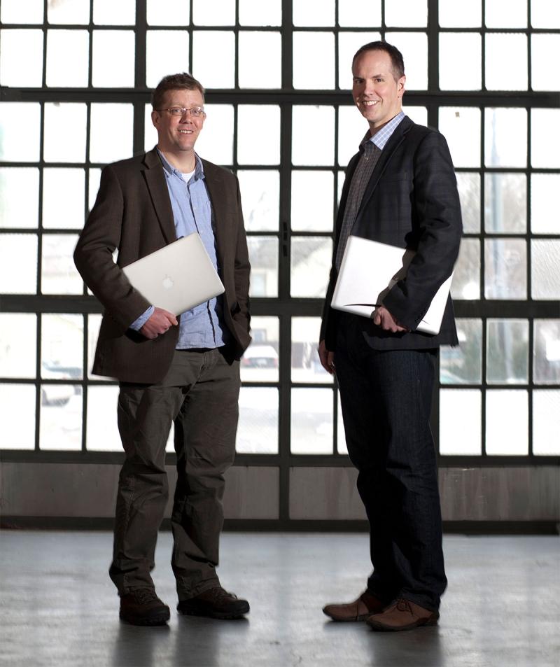 Dan and Tim holding laptops in front of glass wall