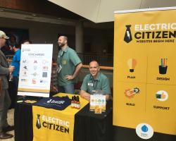 Dan and Adam talking to visitors at the Electric Citizen sponsor table with promotional signage