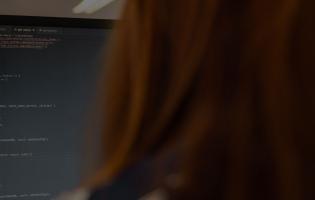 backside view of a person with long red hair looking at a computer screen and code
