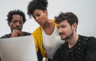 woman in yellow cardigan giving help to two men seated around a computer